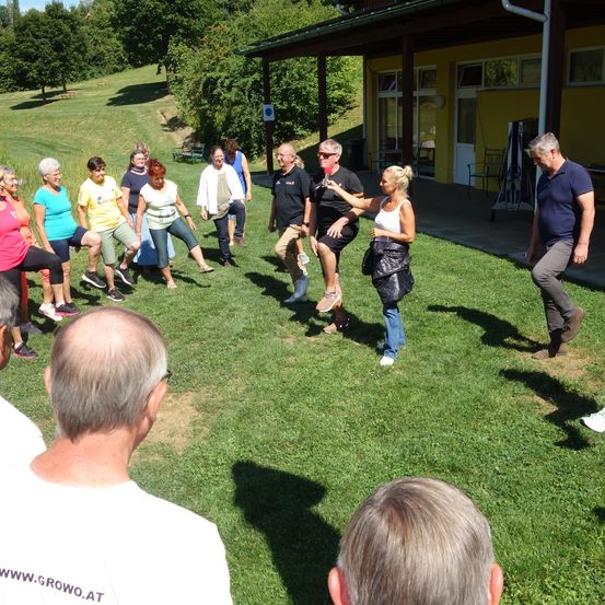 Eine Gruppe von Menschen macht Übungen im Gras, umgeben von einem gelben Gebäude und Bäumen.
