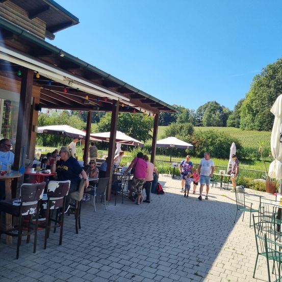 Ein Außenrestaurant mit vielen Gästen, die an einem sonnigen Tag unter Sonnenschirmen sitzen. Bäume und ein Feld sind im Hintergrund zu sehen.