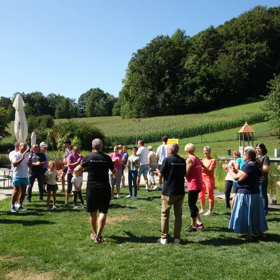 Gruppe von Menschen auf einer Wiese in einem Park. Ein Mann leitet die Gruppe bei einer Übung. Im Hintergrund befinden sich Bäume und ein Feld.