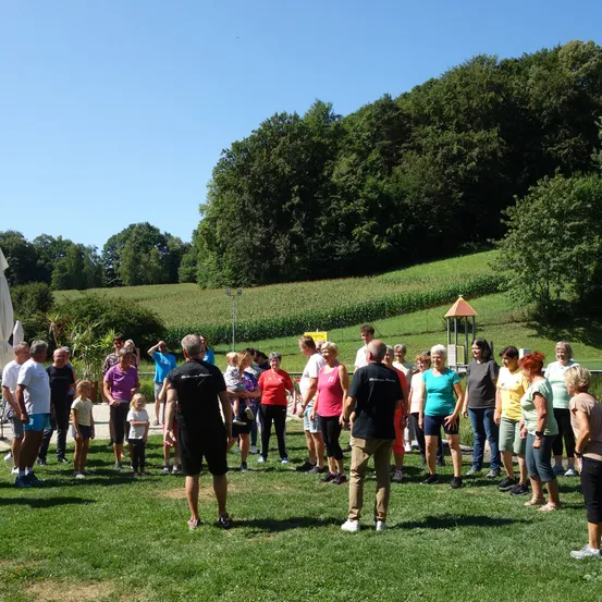 Eine Gruppe von Menschen steht auf einem Rasen unter einem klaren Himmel, umgeben von einem grünen Feld und Bäumen. Einige Personen sind in verschiedenen Farben gekleidet und einige tragen Kinder. Im Hintergrund befindet sich ein Spielhaus.