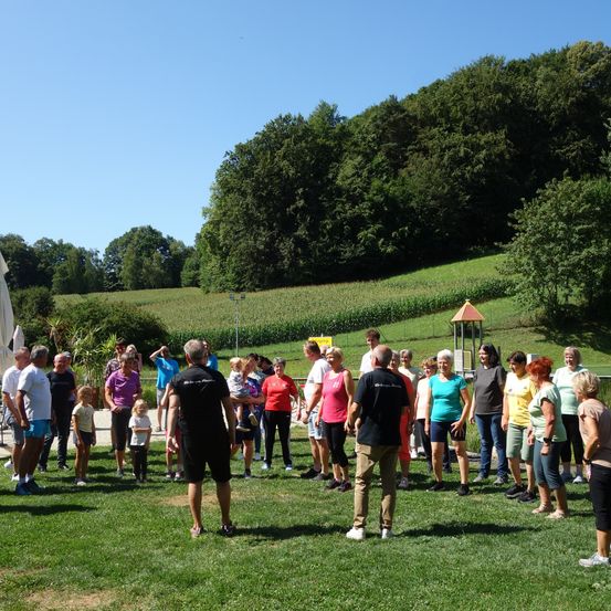 Eine Gruppe von Menschen steht auf einem Rasen unter einem klaren Himmel, umgeben von einem grünen Feld und Bäumen. Einige Personen sind in verschiedenen Farben gekleidet und einige tragen Kinder. Im Hintergrund befindet sich ein Spielhaus.