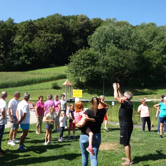 Eine Gruppe von Menschen, darunter Erwachsene und Kinder, versammelt sich auf einer Wiese, möglicherweise für eine Gemeinschaftsveranstaltung. Im Hintergrund sind Bäume und ein gelbes Schild zu sehen.
