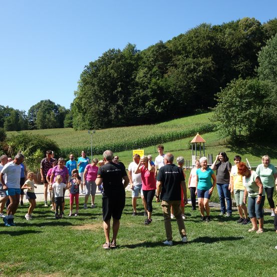 Eine Gruppe von Menschen, einschließlich Erwachsener und Kindern, versammelt sich auf einem Grasfeld, möglicherweise für eine Gemeinschaftsveranstaltung. Sie stehen und einige gehen, möglicherweise an einer Gruppenaktivität teilnehmend. Es gibt Bäume und ein Feld im Hintergrund.