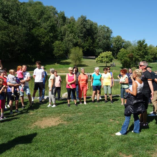 Eine Gruppe von Menschen in lässiger Kleidung steht auf einem Rasenfeld, möglicherweise an einer Gemeinschaftsveranstaltung beteiligt. Die Frau vorne hält eine Plastiktüte. Im Hintergrund befinden sich Bäume und eine Bank.
