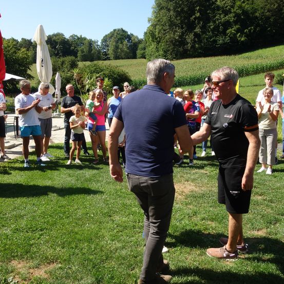 Eine Gruppe von Menschen versammelt sich auf einer Wiese, wobei ein Mann in einem blauen Hemd und ein anderer in einem schwarzen Hemd einander gegenüberstehen. Hinter ihnen stehen und klatschen Menschen, während einige Kinder spielen. Im Hintergrund befinden sich eine grasbewachsene Anhöhe und Bäume.