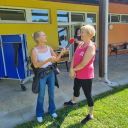 Zwei Frauen sind draußen, eine hält ein Mikrofon und interviewt die andere. Dahinter steht ein gelbes Gebäude mit Glasfenstern. Eine Frau steht drinnen in der Nähe der offenen Tür.