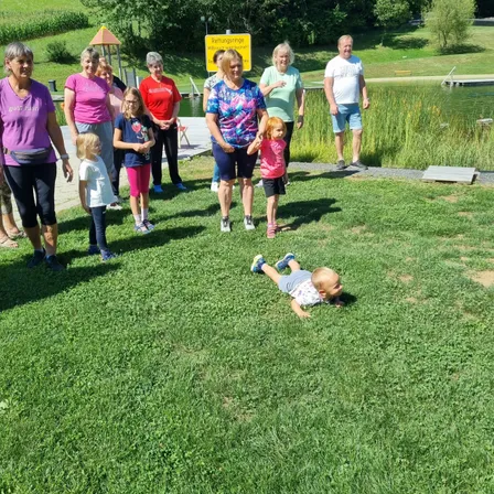 Eine Gruppe von Erwachsenen und Kindern versammelt sich auf einer Rasenfläche in der Nähe eines Teichs. Ein Baby liegt auf dem Gras. Sie sind alle lässig gekleidet.