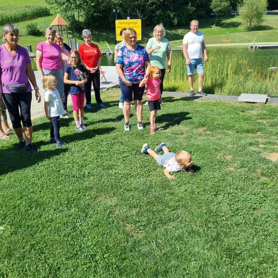 Eine Gruppe von Erwachsenen und Kindern versammelt sich auf einer Rasenfläche in der Nähe eines Teichs. Ein Baby liegt auf dem Gras. Sie sind alle lässig gekleidet.