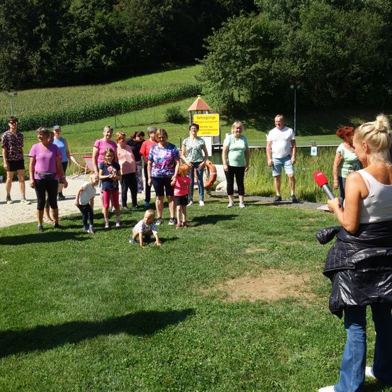 Eine Gruppe von Menschen, einschließlich Erwachsener und Kindern, steht auf einem Rasenfeld. Eine Frau mit einem Mikrofon spricht zu ihnen. In der Nähe gibt es ein gelbes Schild und einen Schwimmring.