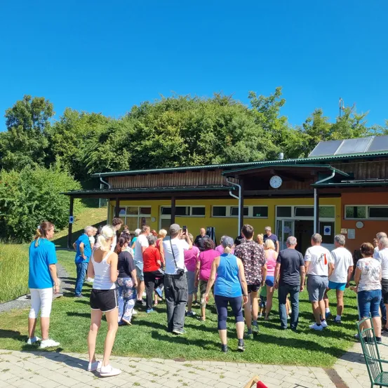 Eine Gruppe von Menschen versammelt sich vor einem Gebäude mit Solarmodulen an einem sonnigen Tag. Einige tragen Turnschuhe, und eine Frau in einem blauen Shirt hält ein Telefon.