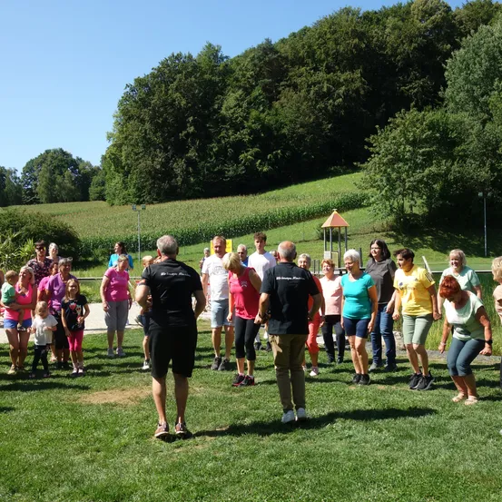 Eine Gruppe von Menschen in verschiedenen Farben steht auf einem Rasenfeld bei klarem Himmel. Bäume und ein Maisfeld sind im Hintergrund zu sehen. Sie scheinen an einer Aktivität teilzunehmen.