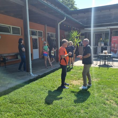 Eine Gruppe von Menschen steht auf einem grünen Rasen vor einem Gebäude. Zwei Männer unterhalten sich, während eine Frau mit einem Baby in der Nähe ist. Dahinter steht eine Frau und beobachtet.