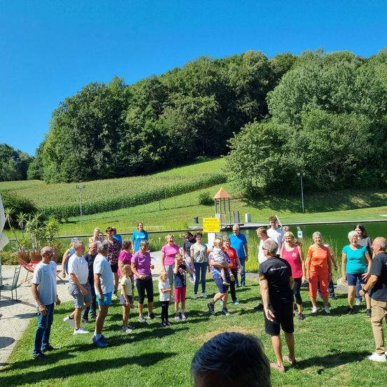Eine Gruppe von Menschen steht auf einem Rasenfeld, wahrscheinlich an einer Veranstaltung teilnehmend. Sie tragen verschiedene Farben von Shirts und Shorts. Hinter ihnen befindet sich ein Gewässer und ein begrünter Hügel mit Bäumen.