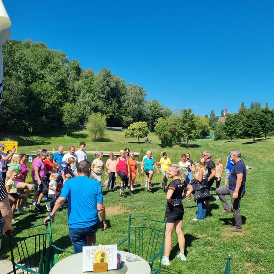 Eine Gruppe von Menschen, darunter Erwachsene und Kinder, versammelt sich auf einem Rasenfeld. Einige stehen, andere sitzen. Eine Frau leitet eine Aktivität. In der Mitte steht ein Tisch mit einem Schild. Bäume und ein Gebäude sind in der Ferne.