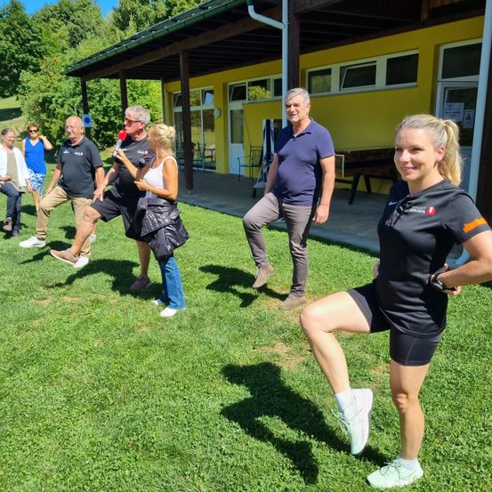Eine Gruppe von Erwachsenen macht draußen auf einem grünen Rasen Sport. Eine Frau hält ein Mikrofon und beugt die Knie. Eine andere Frau trägt einen schwarzen Rucksack. Hinter ihnen steht ein gelbes Gebäude.