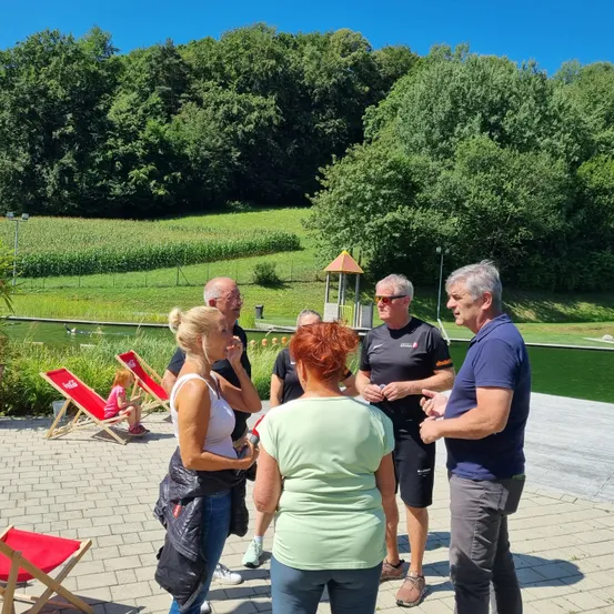 Eine Gruppe von Menschen trifft sich in einem Park und führt ein Gespräch. Ein kleines Kind sitzt auf einem Liegestuhl im Hintergrund. Die Umgebung umfasst Bäume, einen Teich und ein saftiges Feld.