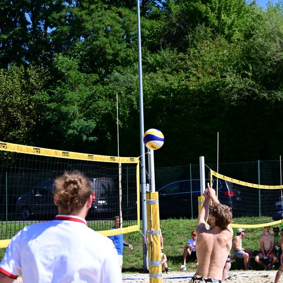 Eine Gruppe junger Männer spielt Beachvolleyball auf einem Feld mit geparkten Autos dahinter. Ein Mann greift nach dem Ball über dem Netz.