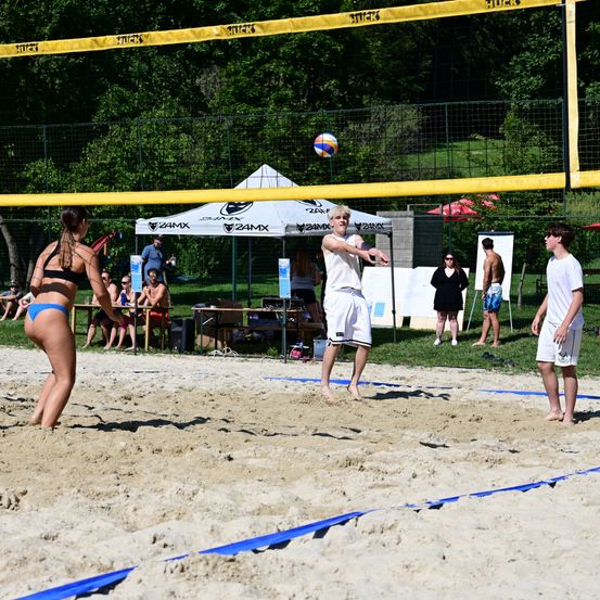 Zwei Personen spielen Beachvolleyball auf einem sandigen Platz. Ein Mann springt, um den Ball zu schlagen, während eine Frau zusieht. Es gibt Leute, die unter einem Zelt sitzen, und andere stehen im Hintergrund.