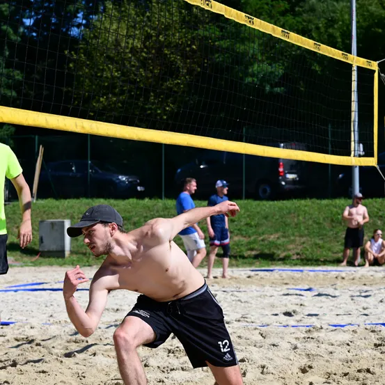 Ein bärtiger Mann spielt Beachvolleyball, rennt auf das Netz zu, während andere im Hintergrund beobachten.