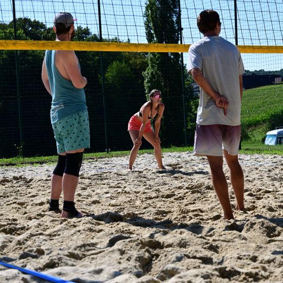 Drei Personen spielen Volleyball auf einem sandigen Platz. Eine Frau in roten Shorts beugt sich nach vorne, möglicherweise um den Ball zu schlagen. Die beiden Männer stehen hinter dem Netz und beobachten das Spiel.