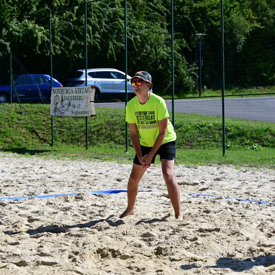 Eine Person in einem gelben T-Shirt und schwarzen Shorts spielt Volleyball auf einem sandigen Platz mit einem Netz. Im Hintergrund befindet sich ein Schild mit Text und Bild, ein Zaun und geparkte Autos.
