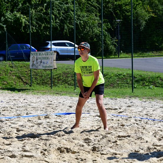 Eine Person in einem gelben T-Shirt und schwarzen Shorts spielt Volleyball auf einem sandigen Platz mit einem Netz. Im Hintergrund befindet sich ein Schild mit Text und Bild, ein Zaun und geparkte Autos.