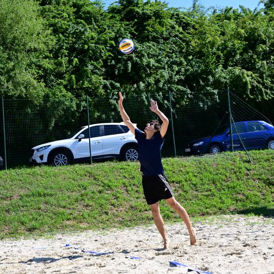 Ein Mann in einem blauen T-Shirt und Shorts spielt Volleyball auf einem Sandstrand. Hinter ihm steht ein Netz. Zwei Autos sind auf einem Grashang in der Nähe des Zauns geparkt.