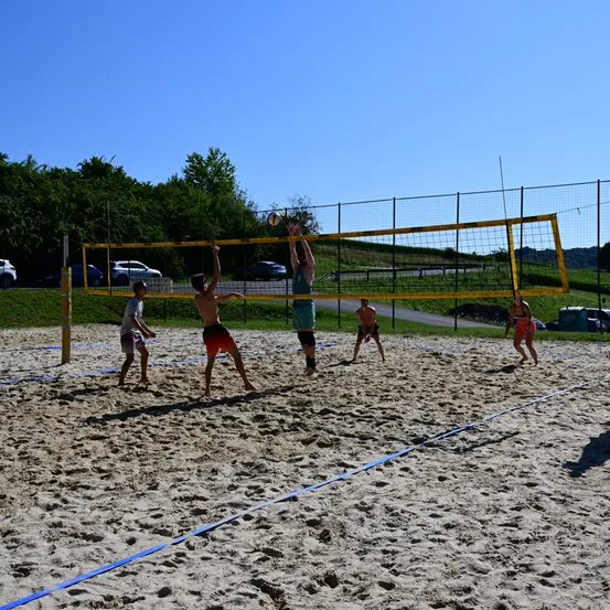 Fünf Personen spielen Volleyball auf einem Sandplatz. Zwei Spieler springen, um den Ball über das Netz zu schlagen. Die anderen beobachten. Der Platz ist von einem gelben Netz umgeben.