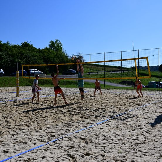 Fünf Personen spielen Volleyball auf einem Sandplatz. Zwei Spieler springen, um den Ball über das Netz zu schlagen. Die anderen beobachten. Der Platz ist von einem gelben Netz umgeben.