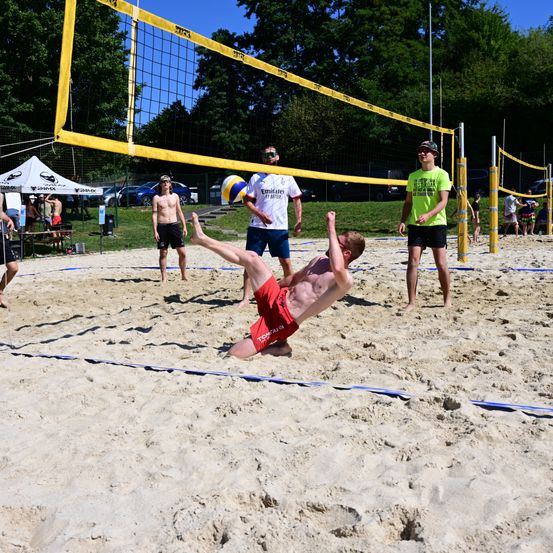 Eine Gruppe von Menschen spielt Volleyball auf einem sandigen Strand. Ein Spieler taucht, um den Ball zu schlagen.