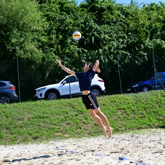Ein Mann springt und versucht, einen Volleyball über ein Netz auf einem Sandstrand zu schlagen, mit geparkten Autos und Grünflächen im Hintergrund.