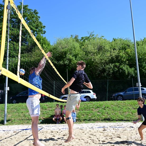 Eine Gruppe von Menschen spielt Volleyball auf einem sandigen Platz mit einem gelben Netz. Zwei Spieler sind am Netz, einer springt zum Angriff, der andere blockt. Zuschauer sitzen an den Rasenseitenlinien, und Autos sind in der Nähe geparkt.