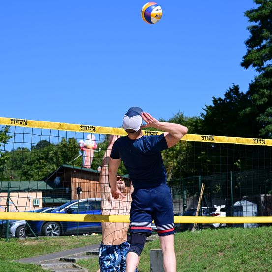 Zwei Männer spielen Volleyball auf einem Freiluftplatz mit einem gelben Netz. Ein Mann versucht, den Ball zu schlagen, während der andere Mann springt, um ihn zu blockieren. Im Hintergrund befinden sich Bäume und ein Gebäude.