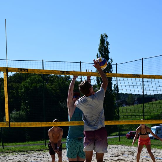 Eine Gruppe spielt Beachvolleyball. Ein Mann springt, um den Ball zu schlagen. Zwei blocken, und eine andere beobachtet. Das Netz ist gelb, und der Boden ist sandig. Bäume und ein Auto sind im Hintergrund.