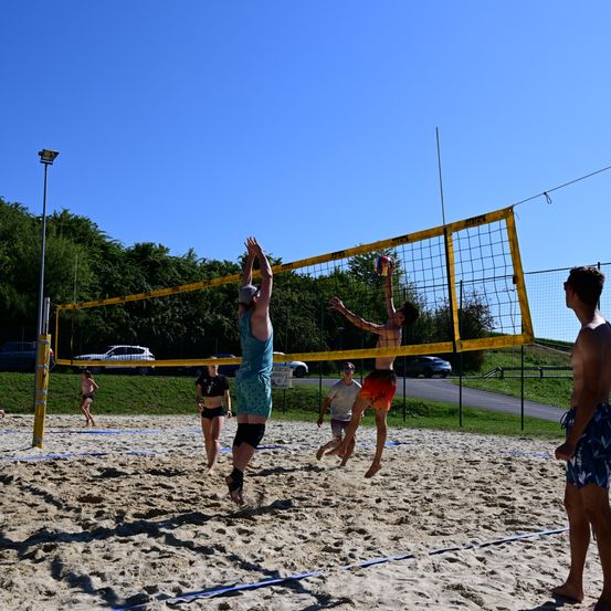 Eine Gruppe von Menschen spielt Volleyball auf einem sandigen Platz mit einem gelben Netz unter einem klaren blauen Himmel.