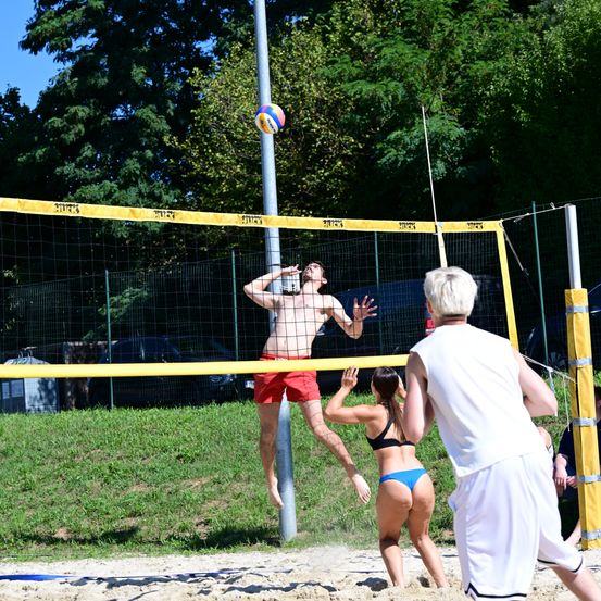 Ein Volleyballspieler springt, um den Ball über das Netz zu schlagen, während ein anderer Spieler versucht zu blocken, an einem sonnigen Strand mit einem Grasbereich und geparkten Autos im Hintergrund.