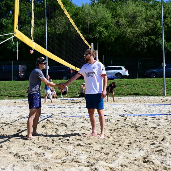 Zwei Männer schütteln sich auf einem Beachvolleyballplatz die Hände. Einer trägt ein graues T-Shirt und blaue Shorts, der andere ein weißes T-Shirt mit Emirates. Zuschauer sitzen auf der Rasenfläche.