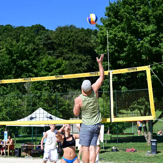Eine Gruppe von Menschen spielt Beachvolleyball im Freien. Ein Mann streckt seine Hand, um den Ball zu schlagen, während eine Frau ihn beobachtet. In der Nähe sind ein Zelt und ein Mülleimer aufgestellt.