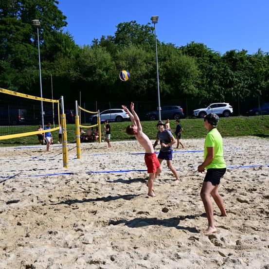 Eine Gruppe von Menschen spielt Volleyball auf einem Sandstrand. Ein Spieler springt, um den Ball zu schlagen.