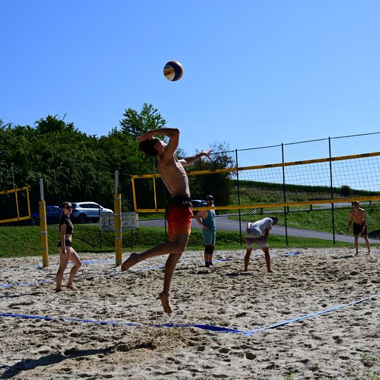 Ein junger Mann springt, um einen Volleyball über das Netz zu schlagen, während andere Spieler zuschauen. Die Umgebung ist ein Outdoor-Volleyballplatz mit einem klaren blauen Himmel.