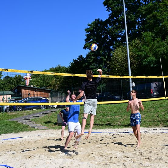 Eine Gruppe von Männern spielt Beachvolleyball auf einem Sandplatz, wobei ein Mann springt, um den Ball zu schlagen.