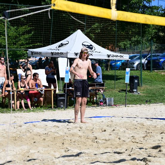 Ein oberkörperfreier Mann steht im Sand eines Beachvolleyballfeldes und hält einen Schläger. Zuschauer sitzen hinter einem Tisch unter einem Zelt mit 24MX-Logo. Bäume und geparkte Autos sind im Hintergrund.