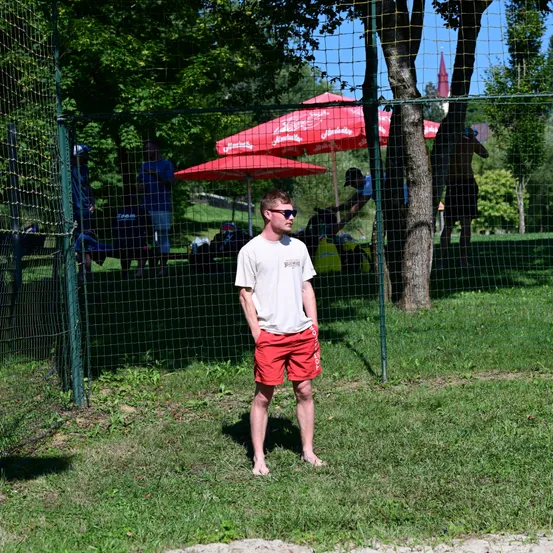 Ein Mann in roten Shorts und einem weißen T-Shirt steht barfuß in einem Grasbereich mit einem Zaun und Bäumen im Hintergrund. Rote Regenschirme stehen hinter dem Zaun.