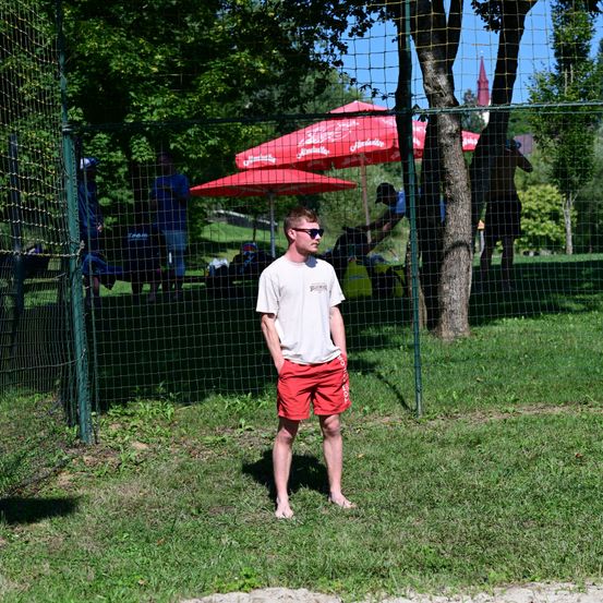 Ein Mann in roten Shorts und einem weißen T-Shirt steht barfuß in einem Grasbereich mit einem Zaun und Bäumen im Hintergrund. Rote Regenschirme stehen hinter dem Zaun.