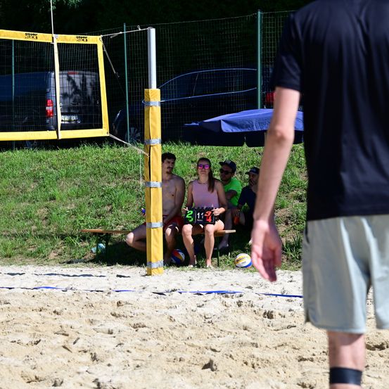 Ein Beachvolleyballspiel mit Spielern auf einer Bank, einer mit einem Trikot mit der Nummer 21134, und einer Person in der Nähe eines Netzes.