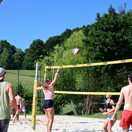 Eine Frau spielt Volleyball am Strand mit einer anderen Frau auf der anderen Seite des Netzes. Es gibt zwei Männer, die zuschauen.
