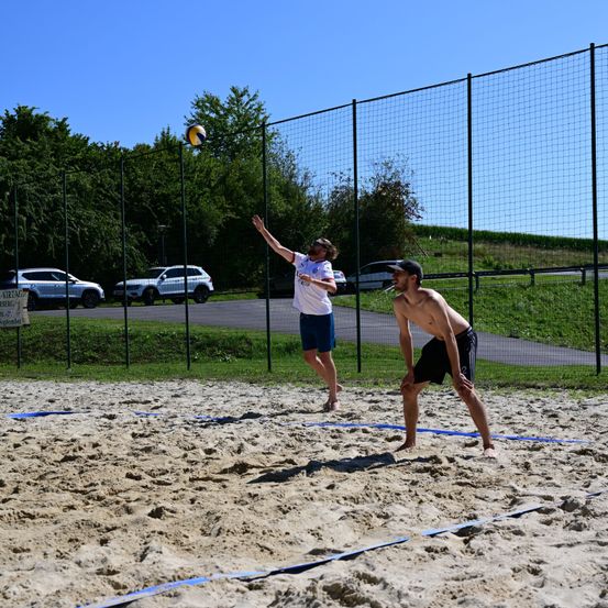 Zwei Männer spielen Volleyball auf einem Sandplatz, einer hebt seine Hände, um den Ball zu schlagen, während der andere sich darauf vorbereitet, ihn zu empfangen. Das Spielfeld ist von einem Netzzaun umgeben, mit Bäumen und geparkten Autos im Hintergrund.