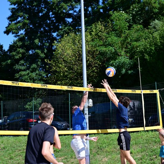 Eine Gruppe von Menschen spielt Volleyball im Freien. Zwei Männer springen, um den Ball zu schlagen. Ein Zaun umgibt das Spielfeld.