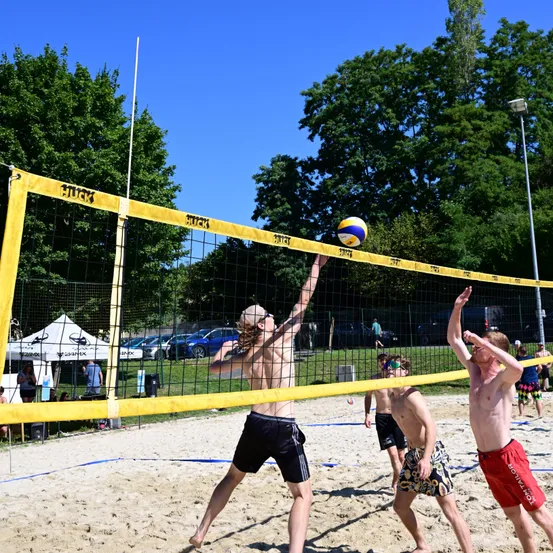 Eine Gruppe von Männern spielt Volleyball auf einem Sandplatz. Ein Spieler springt, um den Ball über das Netz zu schlagen. Die anderen beobachten und sind bereit zu reagieren.