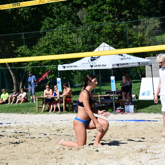 Eine Frau im Bikini spielt Beachvolleyball auf einem Sandplatz. Einige Leute sitzen auf dem Gras hinter dem Spielfeld. Ein Zelt ist im Hintergrund aufgebaut.